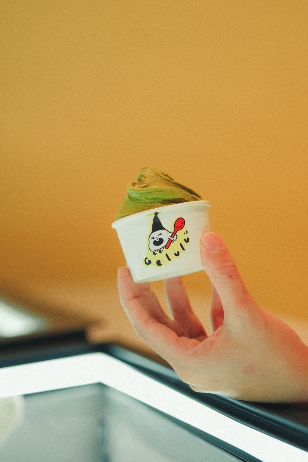 A hand holding a Gelulu cup of matcha and pistachio gelato against the shop's yellow wall